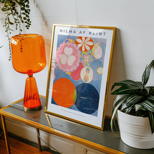 Framed Hilma af Klint artwork on a table with an orange lamp and plant.