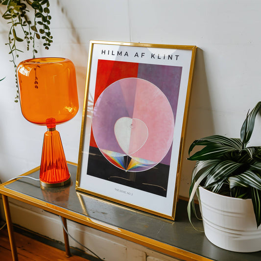 Framed Hilma af Klint artwork on a table with an orange lamp and plant.