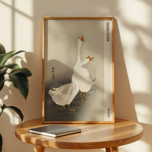 Framed artwork of two geese on a wooden table with a plant in the background. Two Geese by Ohara Koson