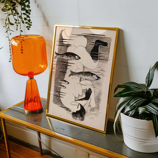 Orange glass lamp on a table with a framed artwork and plant in the background