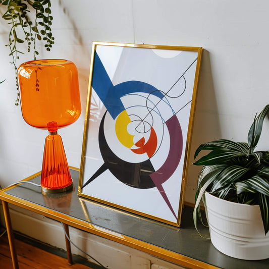 Orange glass table lamp, abstract art print, and potted plant on a wooden surface. Dynamic Construction, Penetration of Spirals and Diagonals by Sophie Taeuber-Arp.