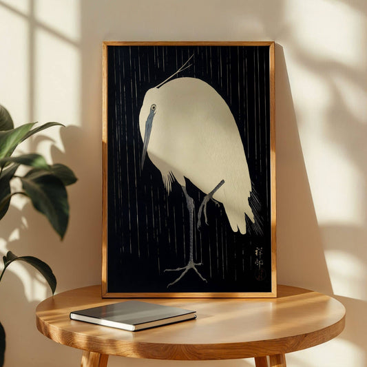 Framed artwork of a bird on a wooden table with a plant in the background. Egret in the Rain by Ohara Koson