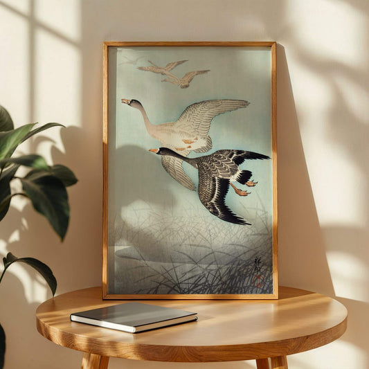 Framed artwork of two birds on a wooden table with a plant in the background. Great Geese in Flight by Ohara Koson