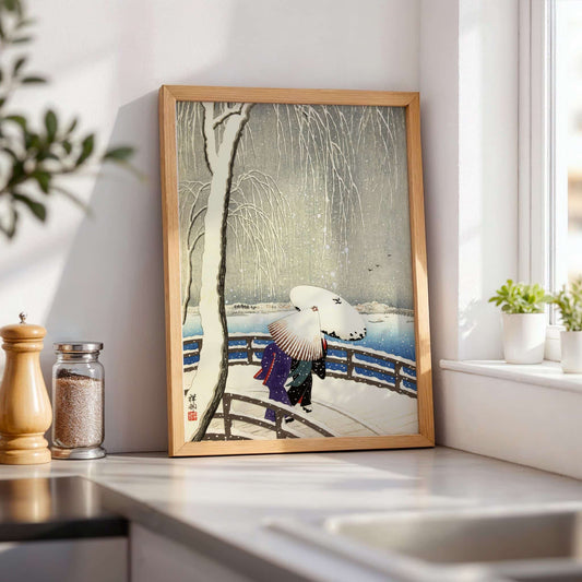 Framed artwork of a snowy scene with people under umbrellas on a wall in a bright room. Snow on Willow Bridge by Ohara Koson