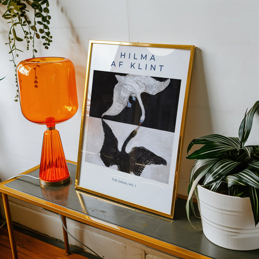 Framed artwork of Hilma af Klint on a table with an orange lamp and plant.
