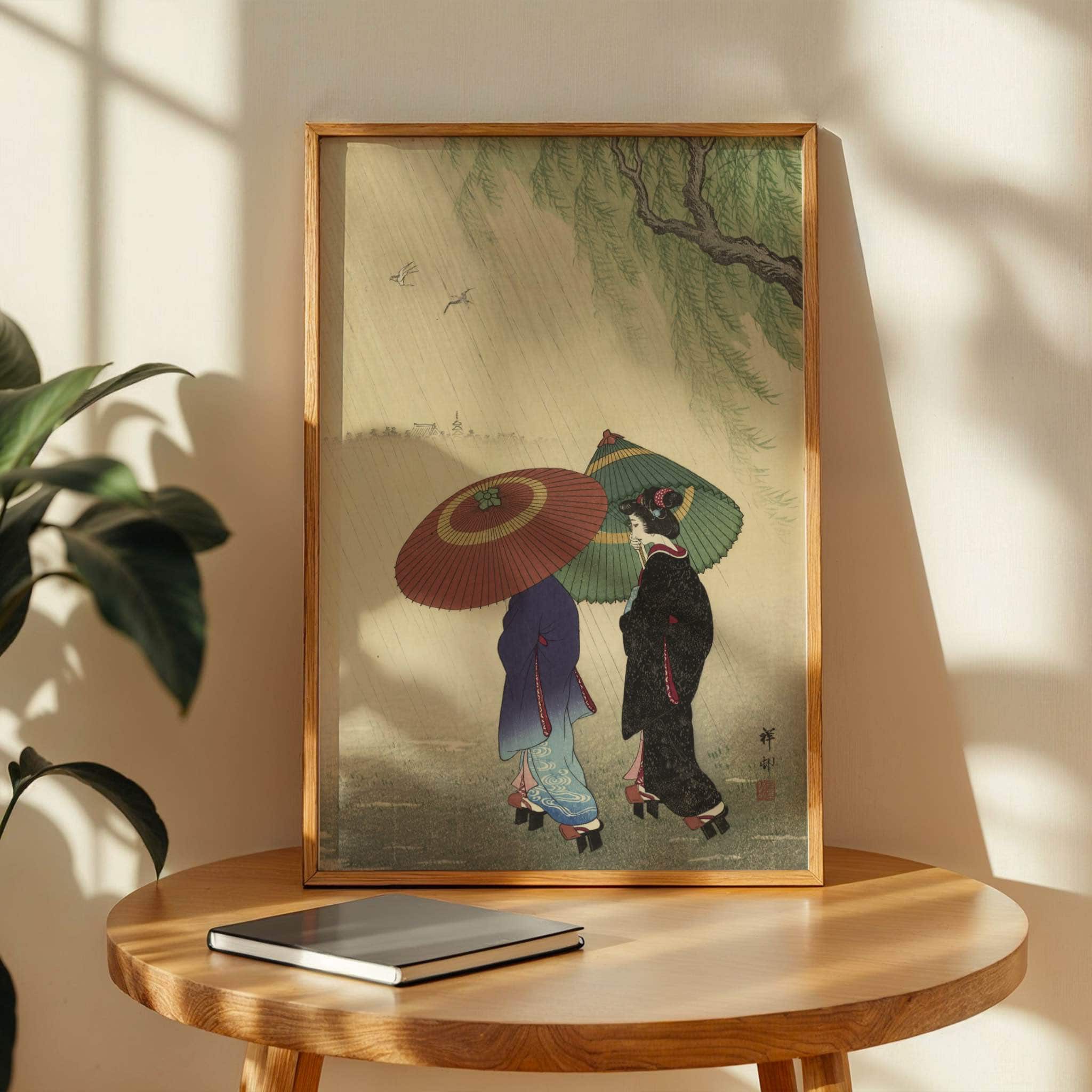 Two Women in the Rain by Ohara Koson. Framed artwork of two people with umbrellas on a wooden table with a plant in the background